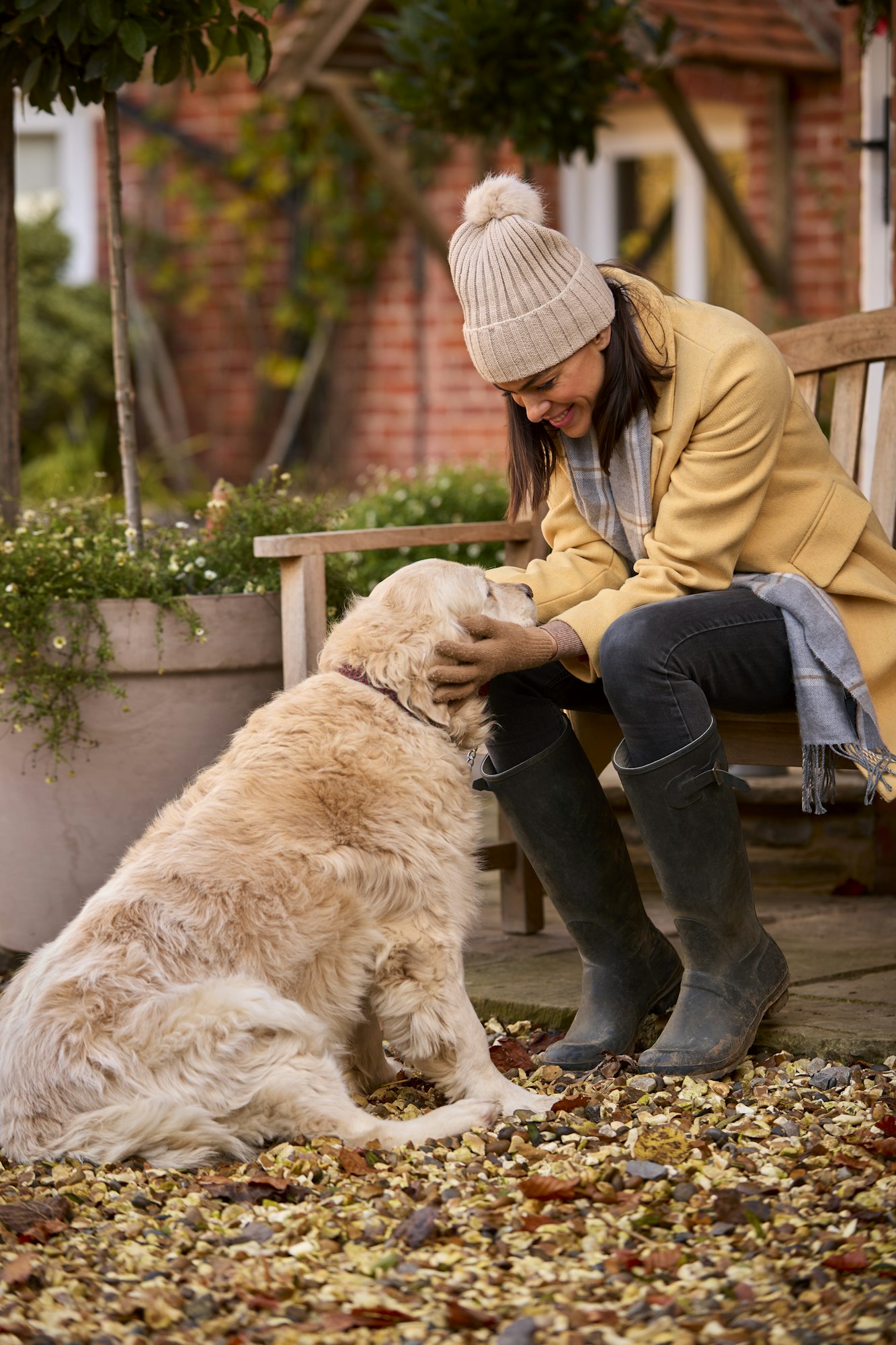 Woman Wearing Hat And Scarf Sitting On Bench Outside House With Pet Dog Ready For On Autumn Walk