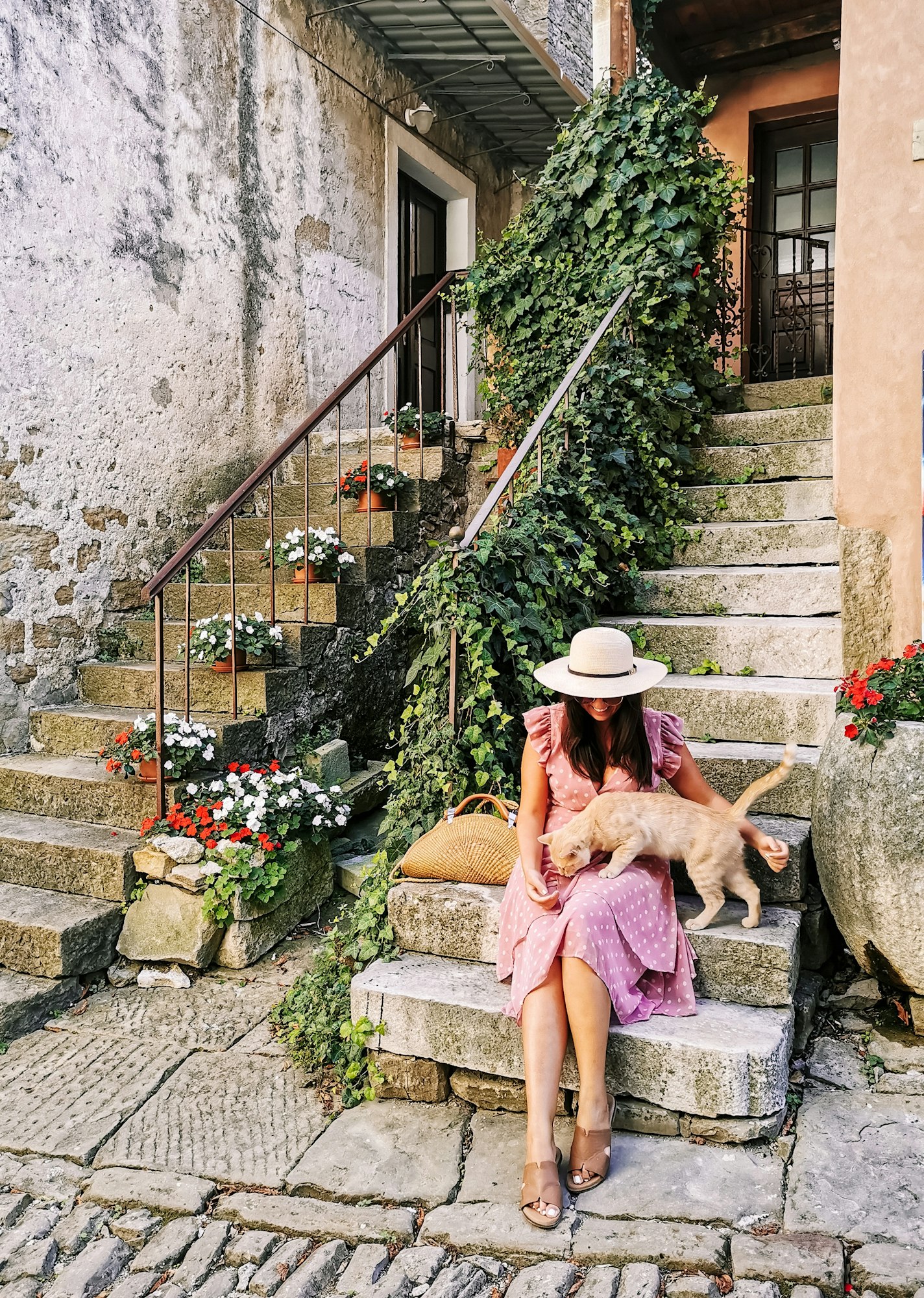 Young woman in pink summer dress petting a cat on stairs in front of old house in idyllic town